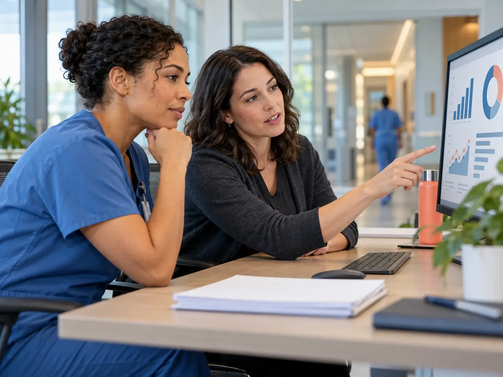 A nurse in scrubs and a colleague reviewing charts on a monitor at a hospital desk, with a corridor behind them.